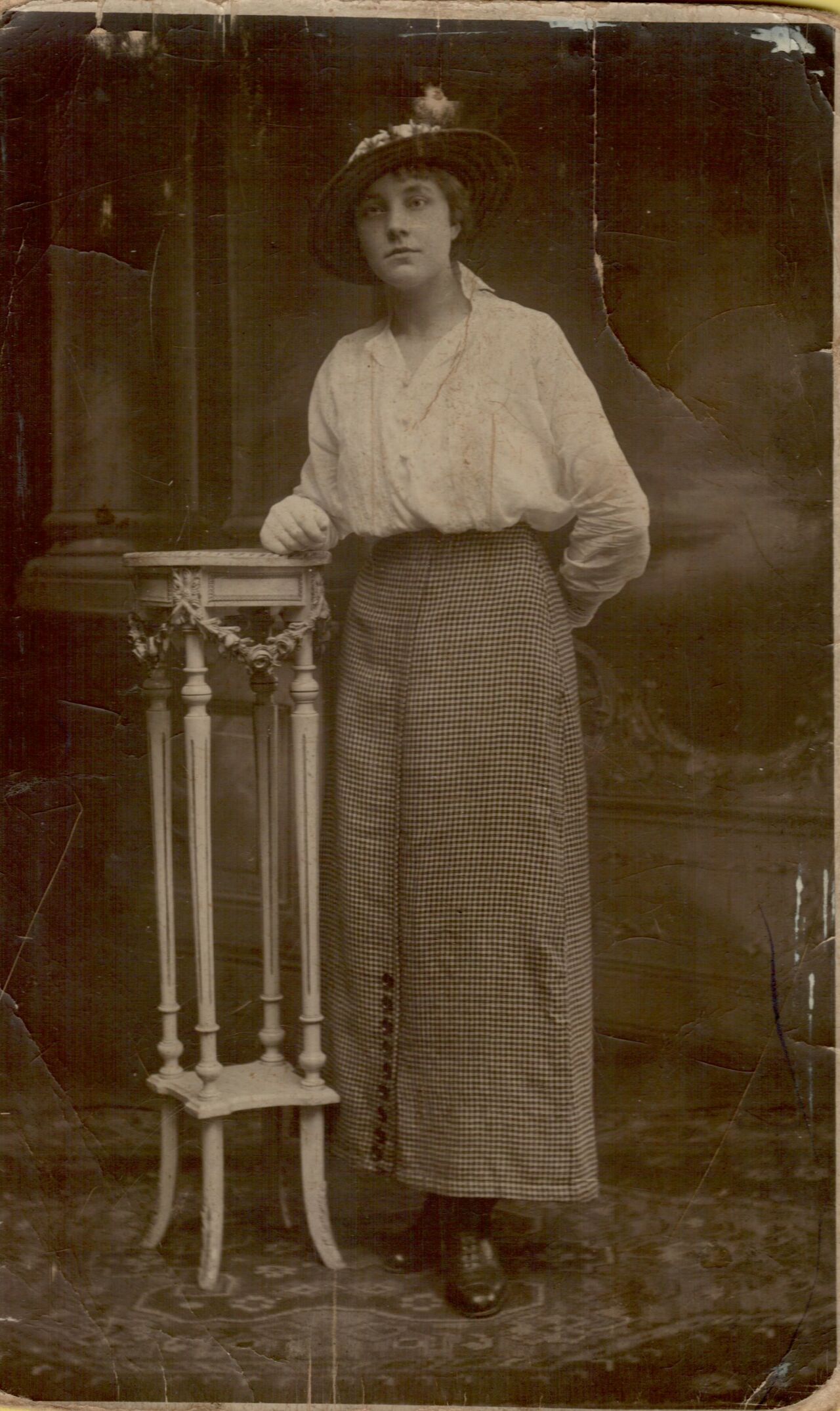 A woman in an old-fashioned outfit stands next to a small decorative table, wearing a hat with a feather.