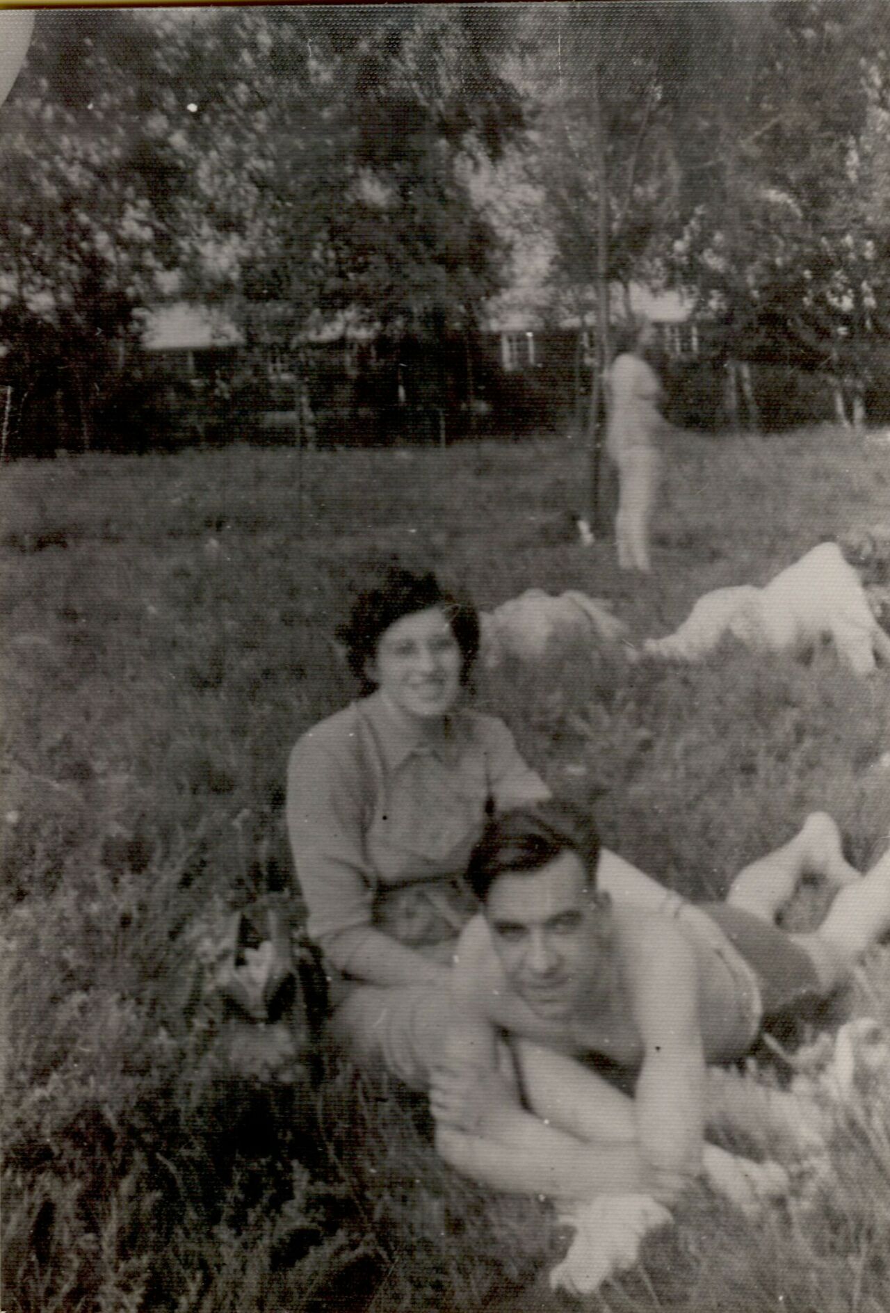 A woman sits on the grass, smiling, while a shirtless man lies in front of her, resting on his arms.