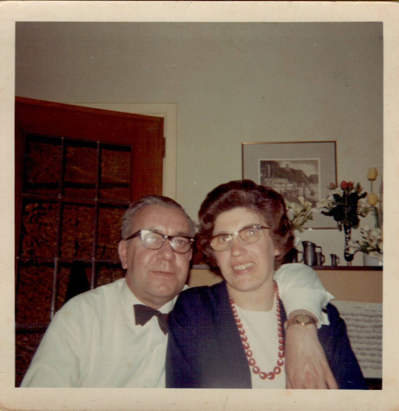 An older couple wearing glasses sits closely together, smiling, with the man's arm around the woman's shoulder.