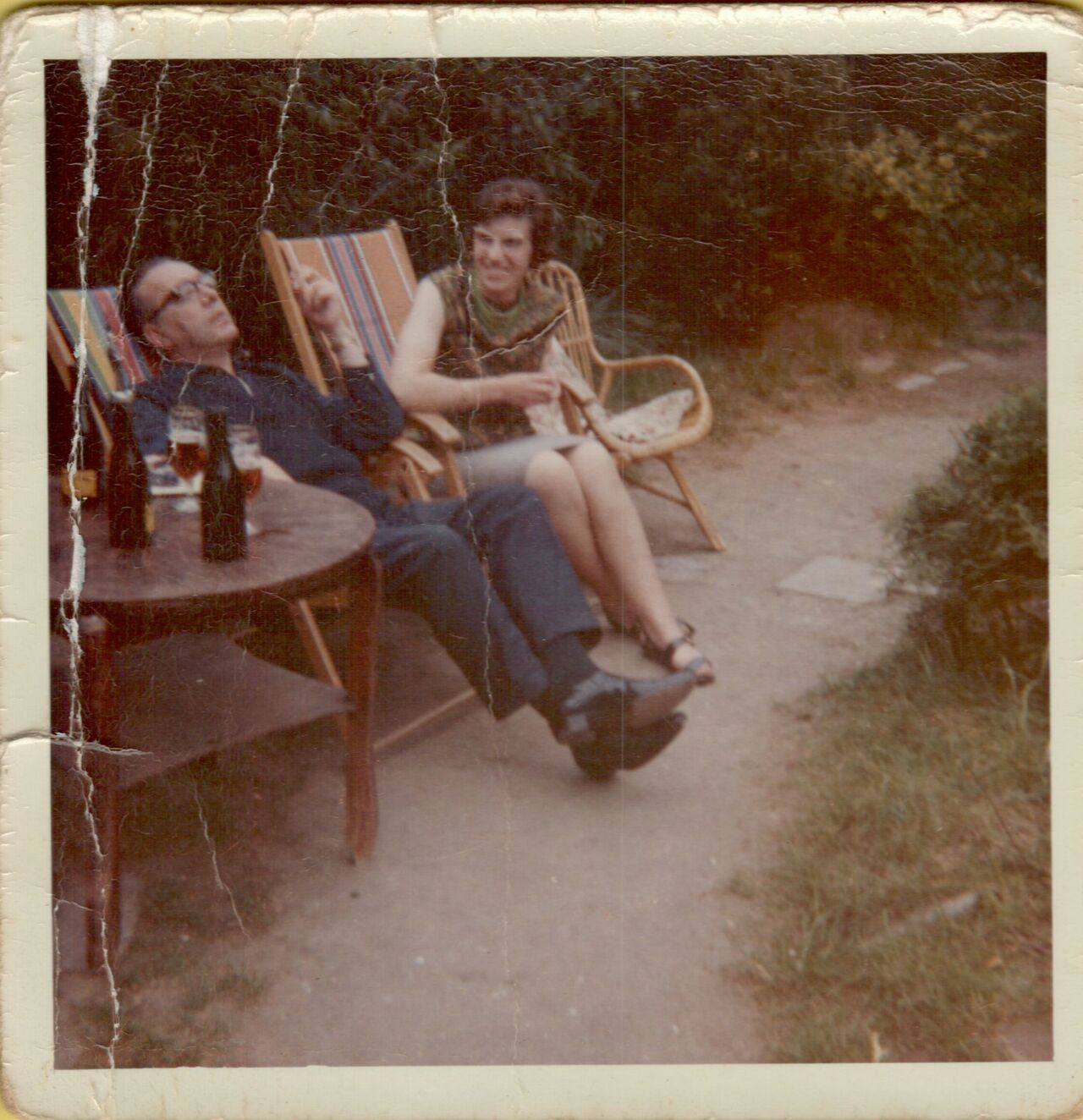 An older man and woman sit outdoors on lounge chairs, talking and relaxing with drinks on a nearby table.