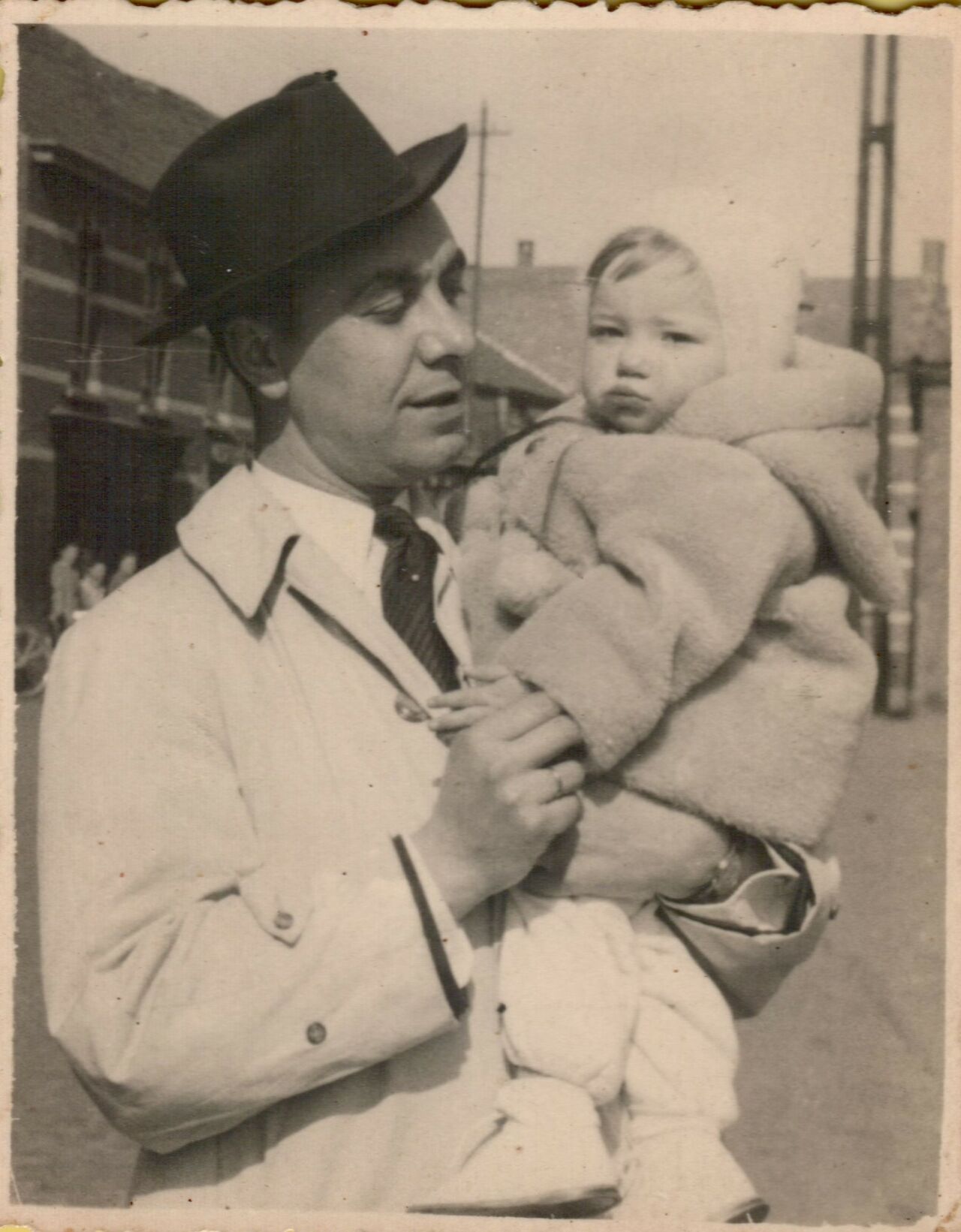 A man wearing a hat and coat holds a warmly dressed baby in his arms outdoors.