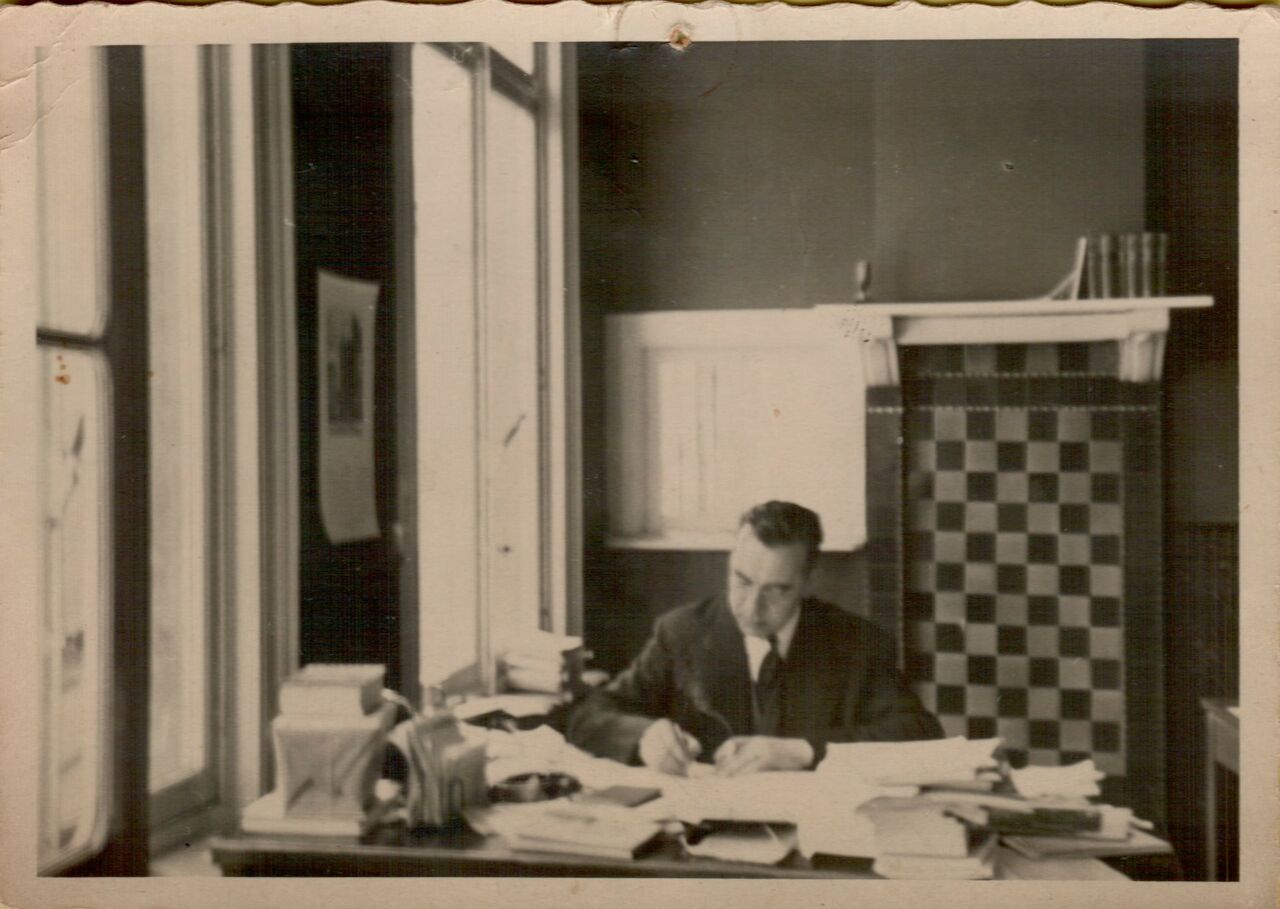 A man in a suit sits at a desk, writing on papers in an office with large windows.