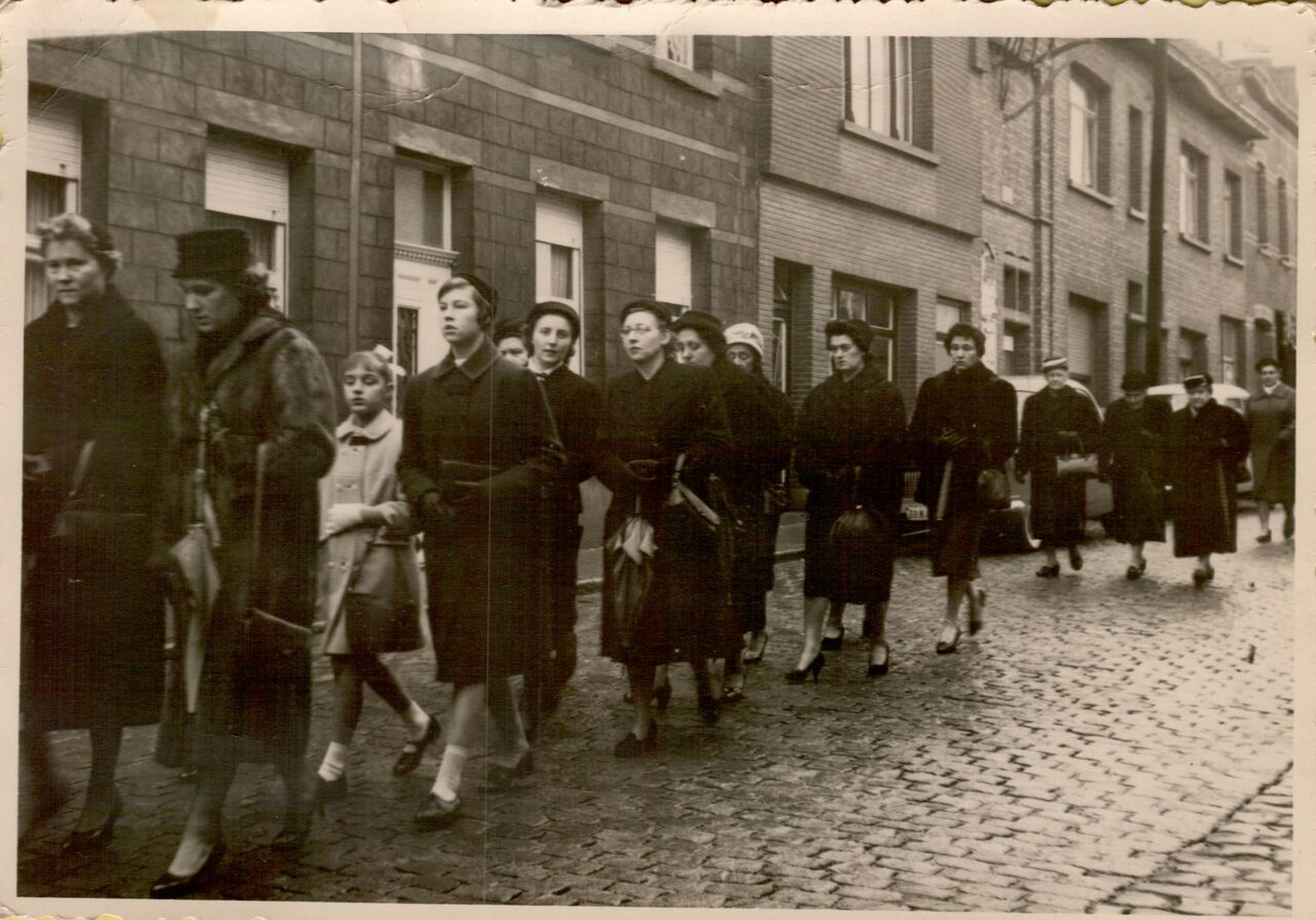 A group of people, mostly women, walk down a cobblestone street wearing coats and carrying bags.