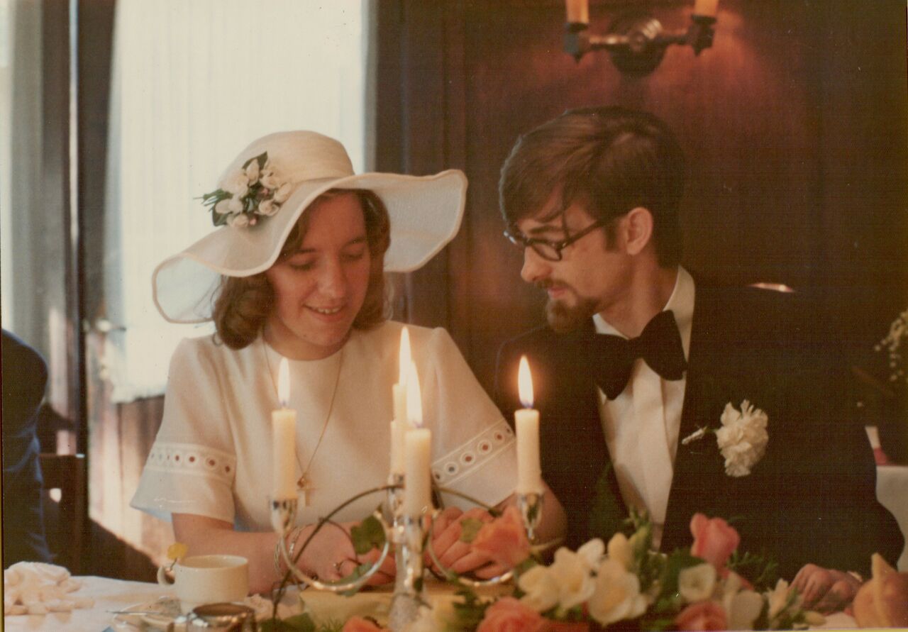 A bride and groom sit at a decorated table with candles, smiling as they cut their wedding cake.