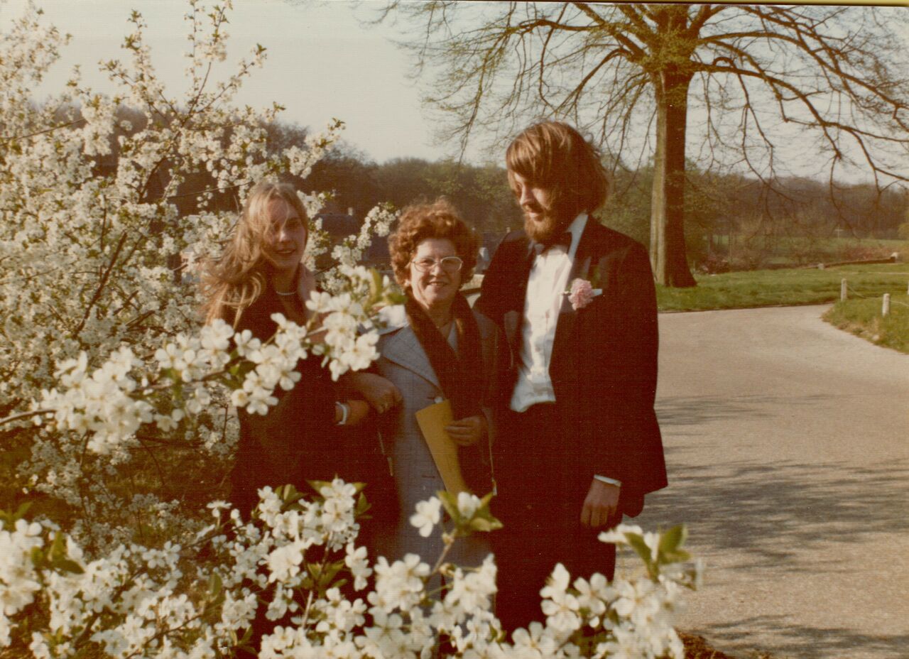 A man in a tuxedo stands with two smiling women, posing near blooming trees on a sunny day.