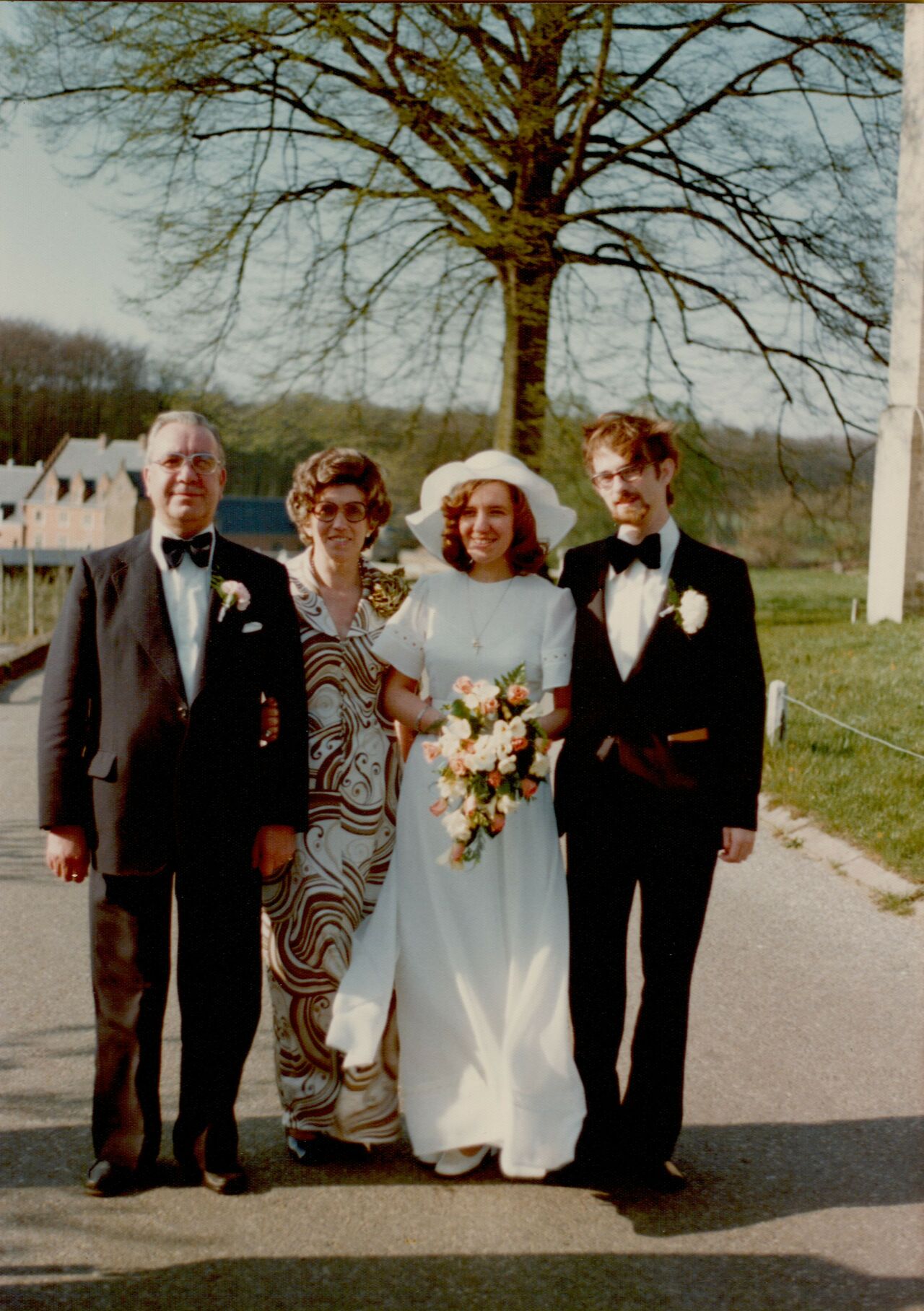 A bride and groom stand outdoors with two older guests, all dressed formally and smiling at the camera.