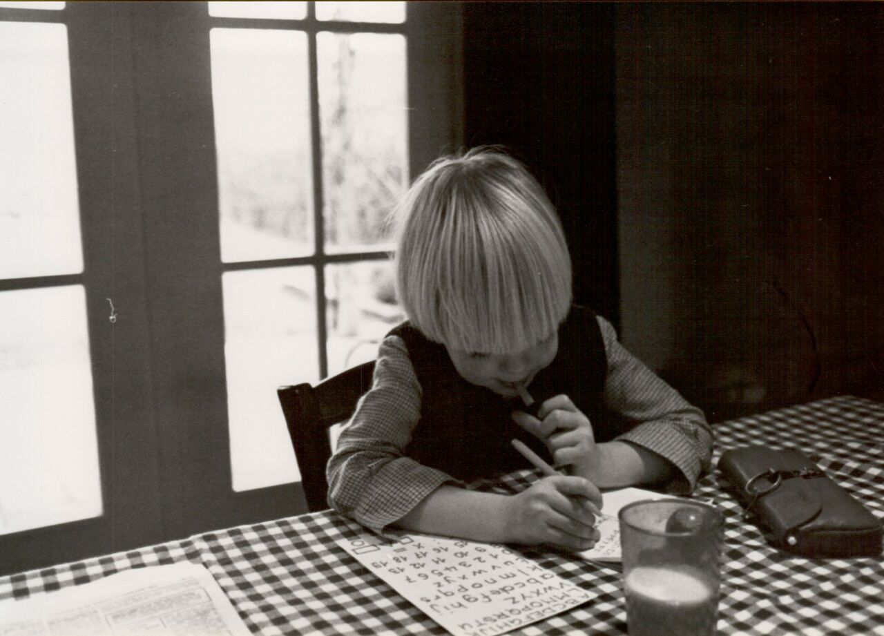 A child sits at a table, concentrating while writing on a worksheet with a pencil.