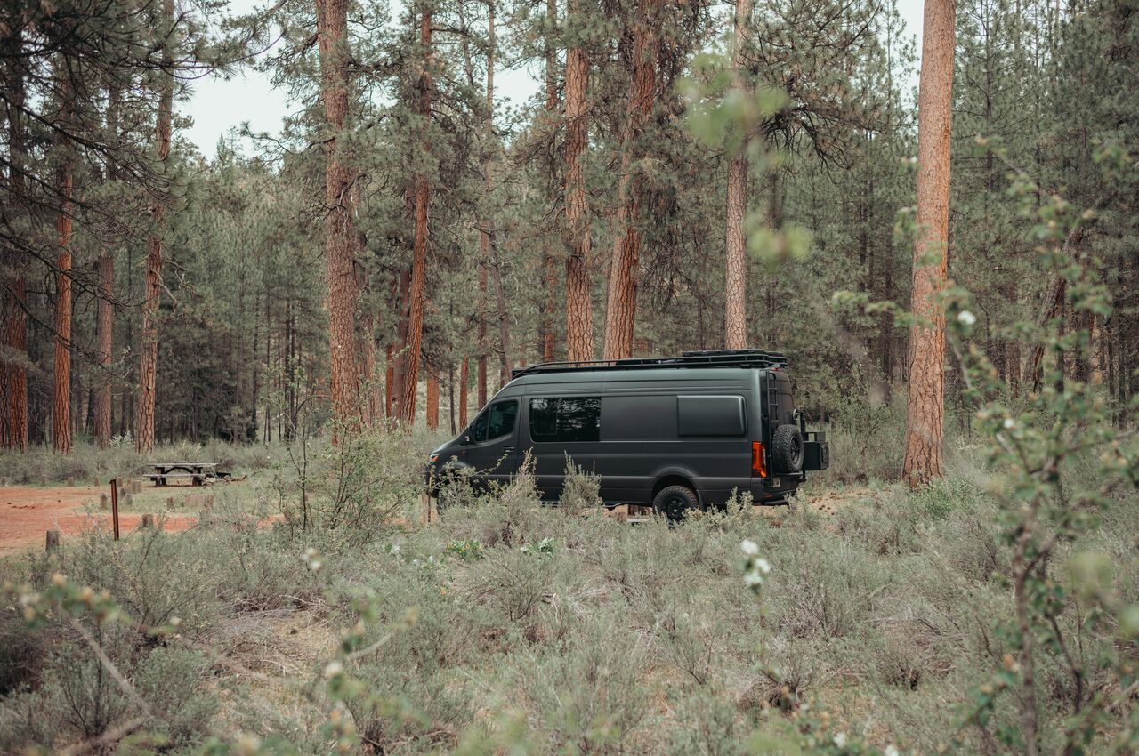 Camp Sherman campground A black camper van is parked in a forested campground surrounded by tall pine trees.