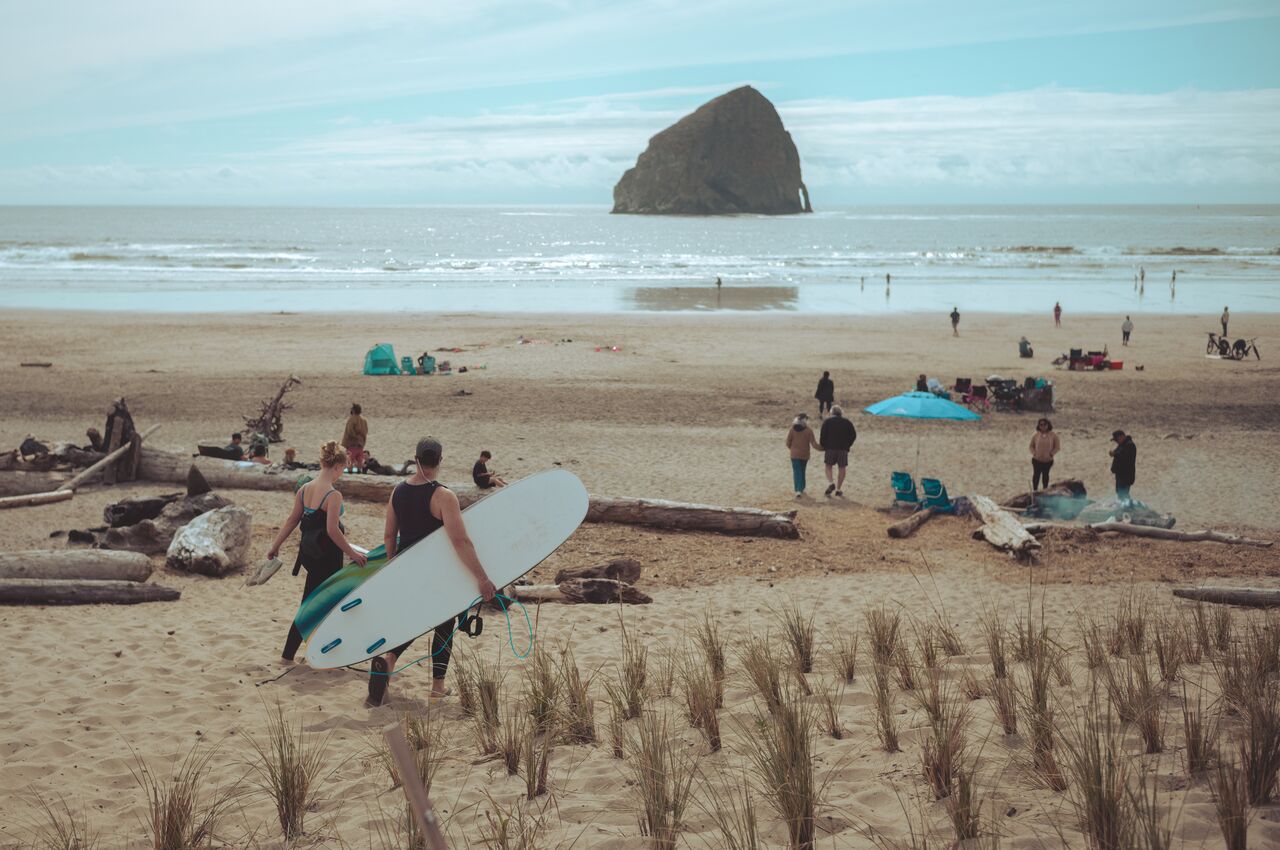 Chief Kiawanda Two people carrying surfboards walk toward the beach with a large rock formation in the water.