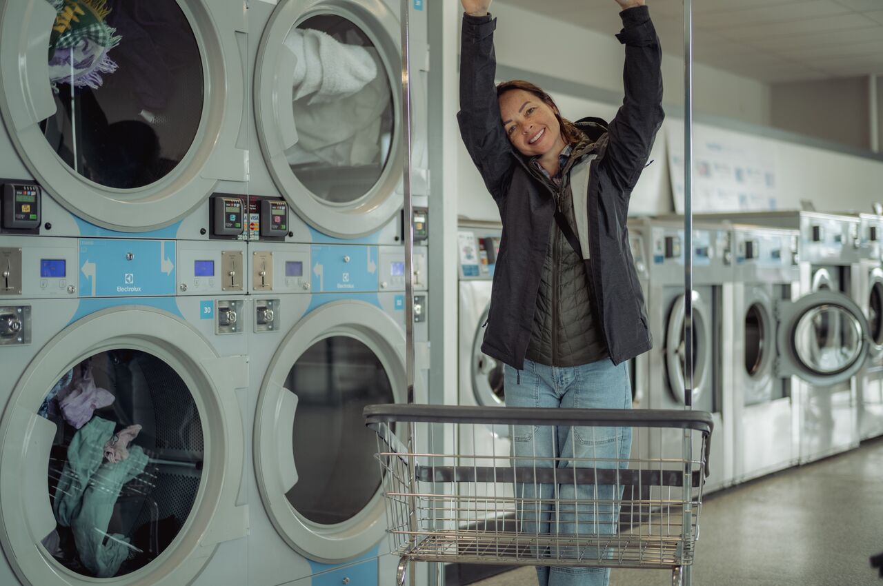 Laundromat Person stretching and smiling next to a laundry cart in front of dryers at a laundromat.