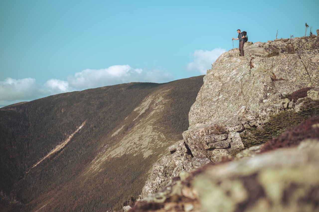 A hiker on top of a cliff, raising his trekking poles in triumph.