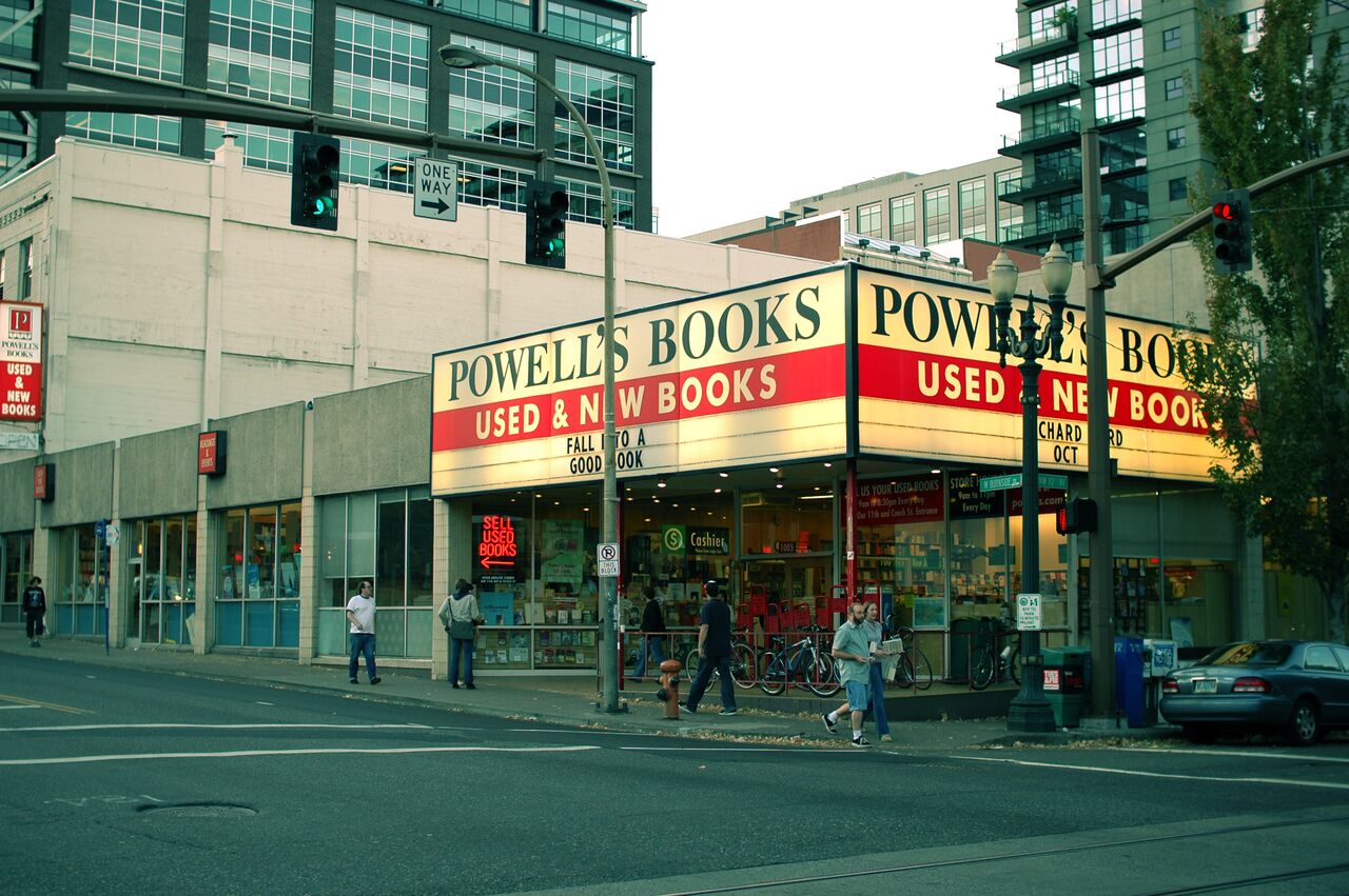 People walk past Powell's Books, a large bookstore with a bright sign advertising used and new books.