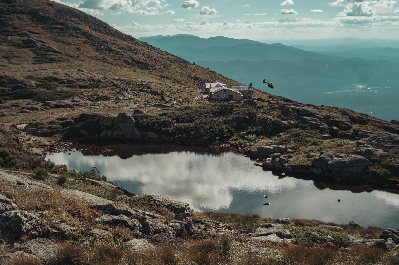 A helicopter flies near a mountain hut and alpine pond, with rocky terrain and hazy ridgelines in the background.