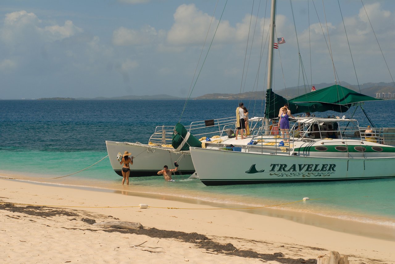 A catamaran named "Traveler" is anchored near a beach while people swim, walk, and relax on board.