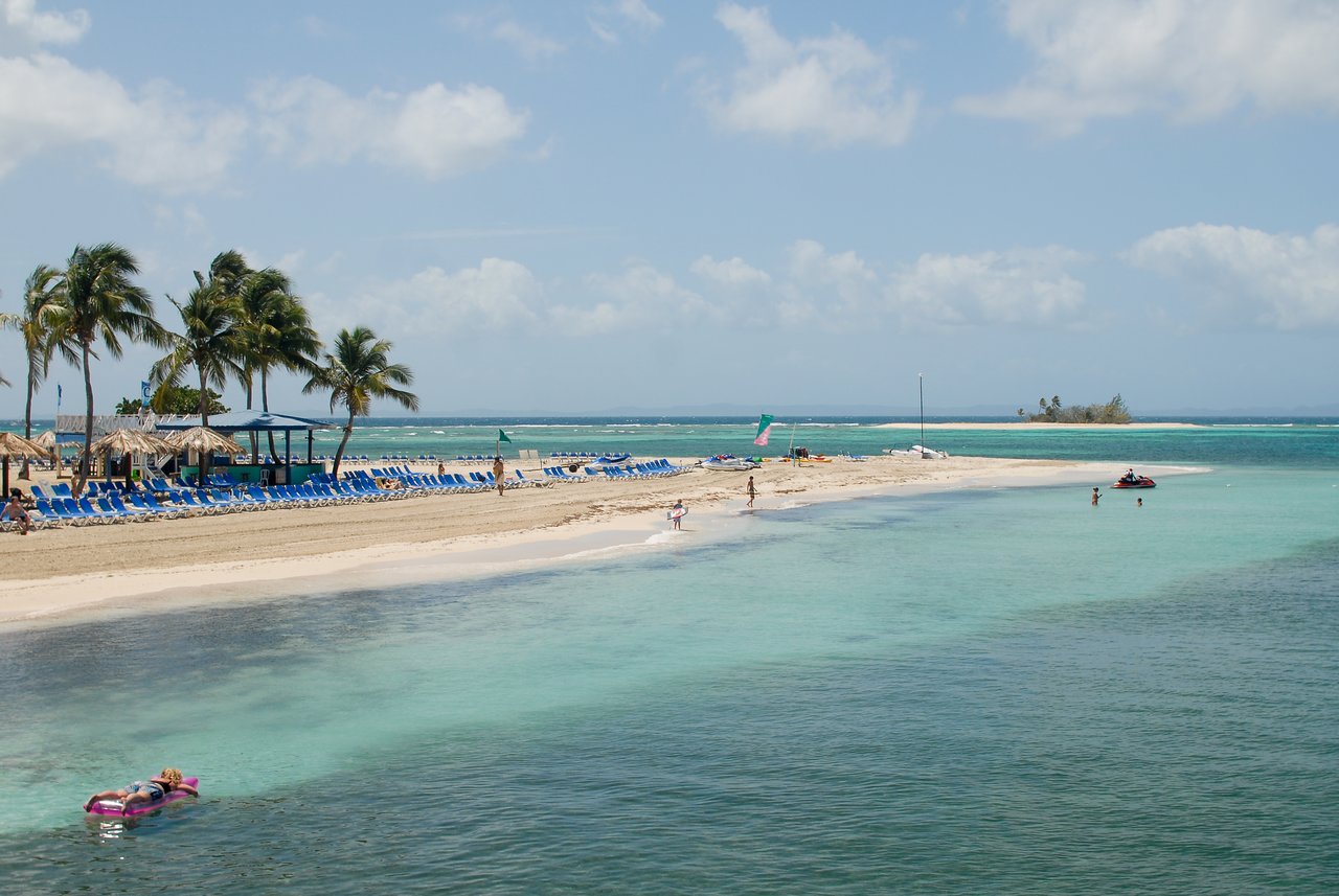 A sandy beach with blue lounge chairs, palm trees, and people swimming or relaxing in the clear water.