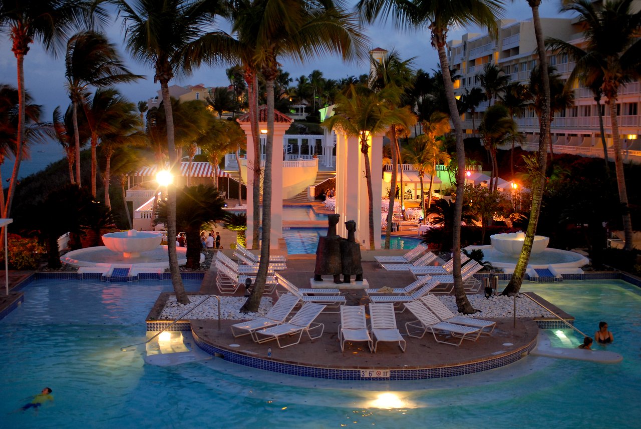 A resort pool area at night with lounge chairs, fountains, and people swimming and relaxing in the water.