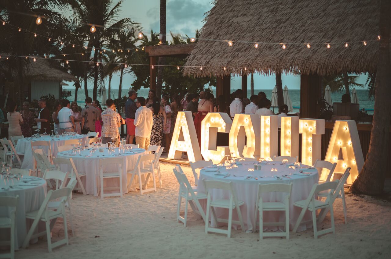 Large illuminated letters spelling &quot;Acquia&quot; are displayed next to tables arranged for a beachside dinner.