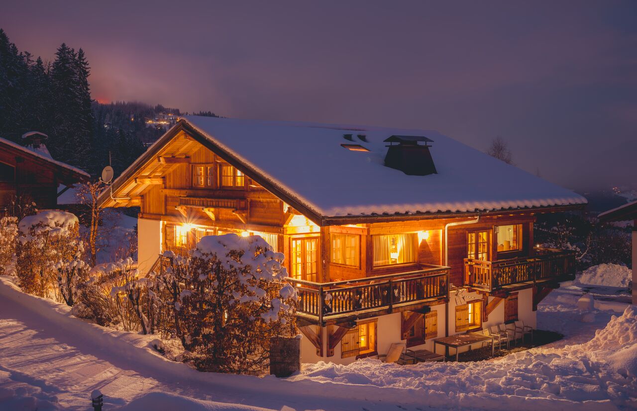 A snow covered wooden alpine chalet glows with warm interior light against a dark purple winter sky.
