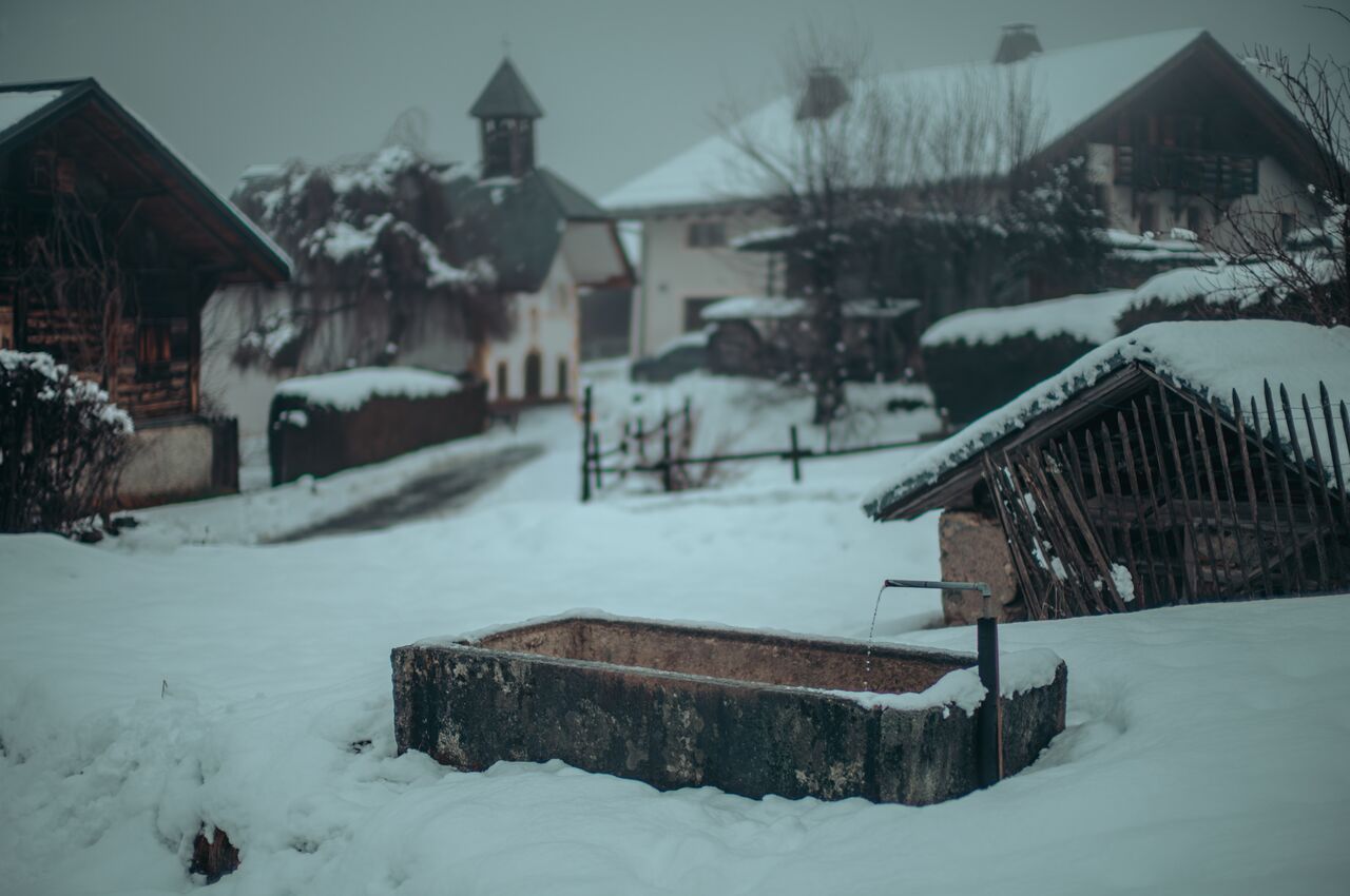 A snow covered stone water trough with a running spout sits in a snowy alpine village.
