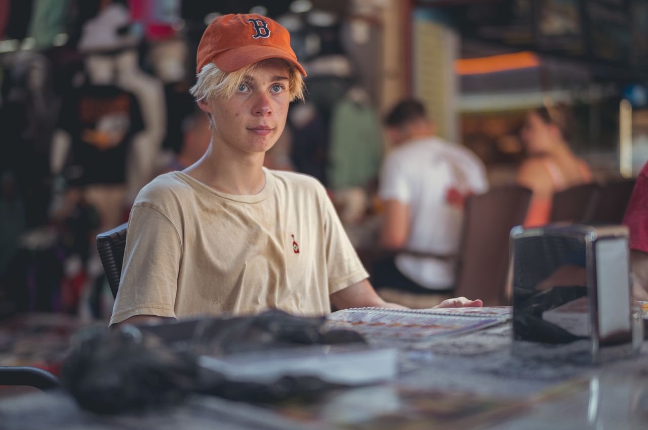 A young adult sits at a restaurant, wearing an orange Boston hat.