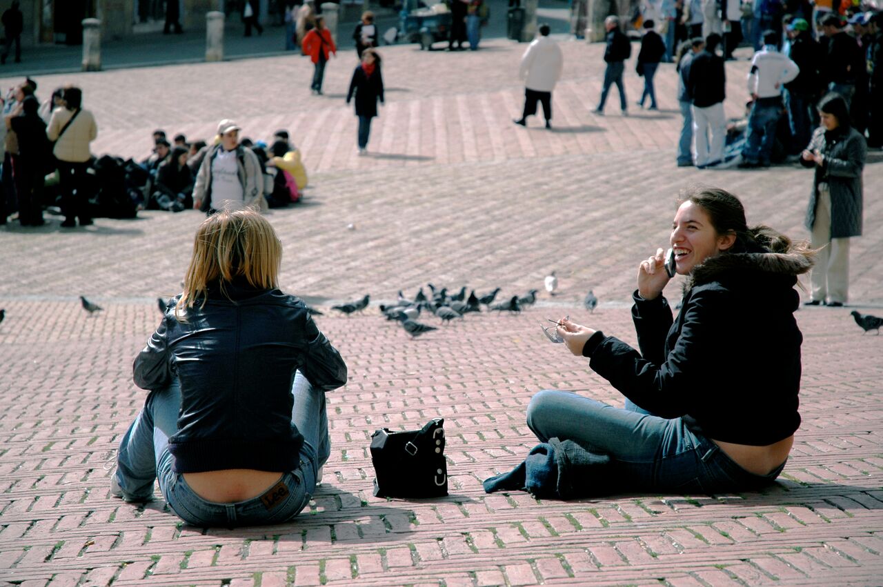 Two young women sit on a brick pavement, one laughing while talking on the phone, with pigeons nearby.