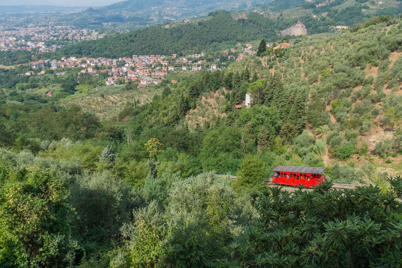 A red funicular railway car travels up a hillside surrounded by green vegetation near Montecatini Alto, Italy.