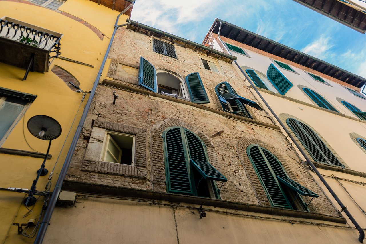 A rustic Italian building with green wooden shutters, open windows, and a brick facade, viewed from below.