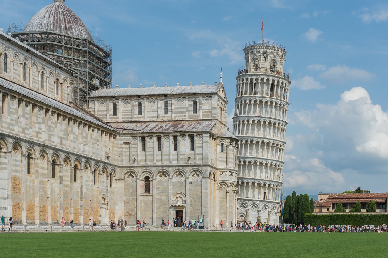 The Leaning Tower of Pisa stands at an angle beside a historic cathedral, with tourists walking nearby.