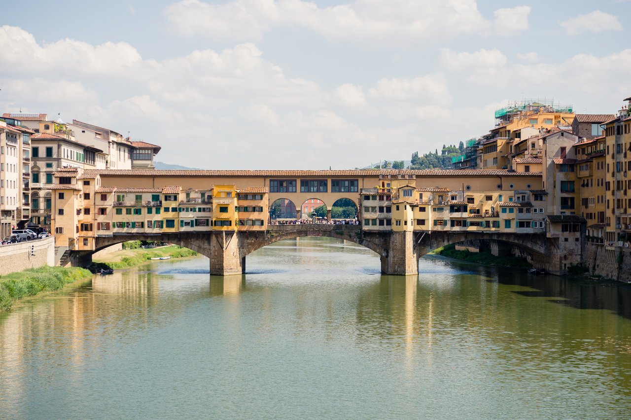 The Ponte Vecchio in Florence, a historic bridge with shops, spans the Arno River under a partly cloudy sky.