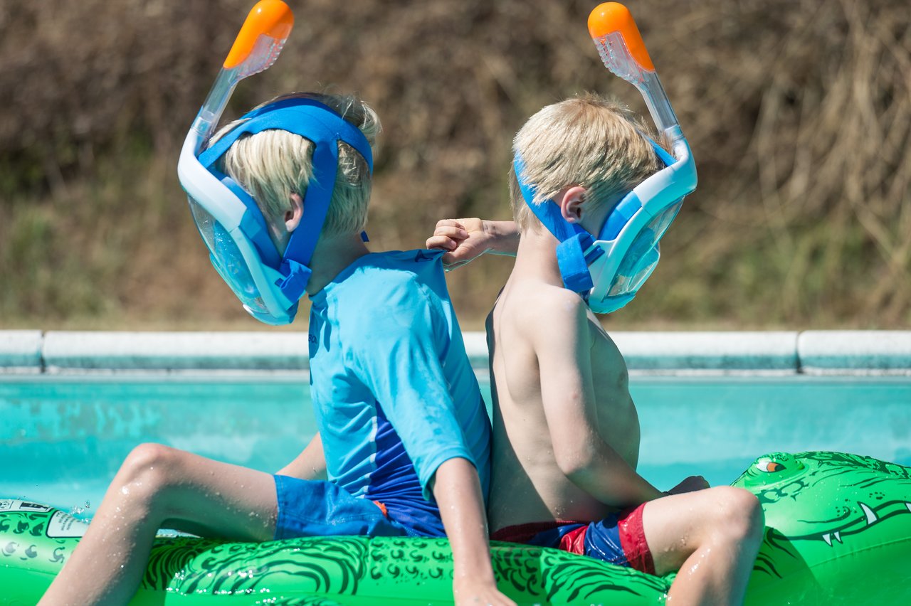 Two children wearing snorkel masks sit back-to-back on a green inflatable float in a swimming pool.