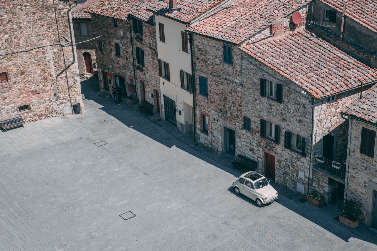 A small vintage car is parked on a stone-paved square beside old brick buildings with wooden shutters.