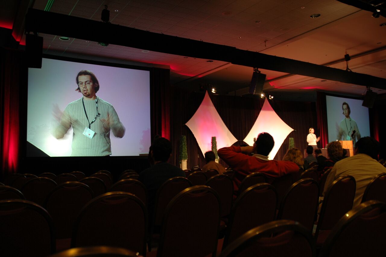 A presenter speaks on stage at a conference, with a large screen displaying a close-up of him gesturing.