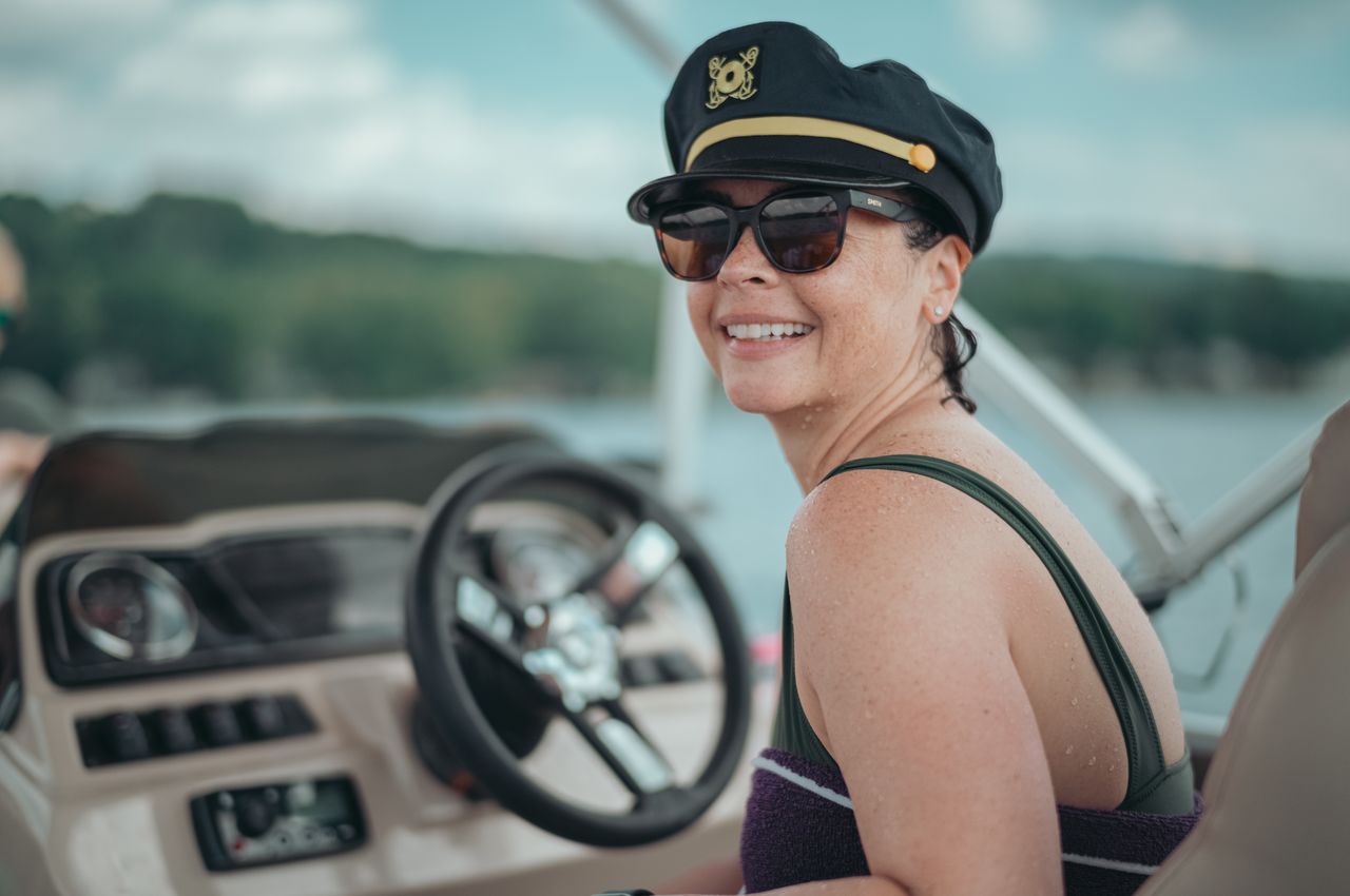 A woman wearing a captain's hat seated at the steering wheel of a boat.