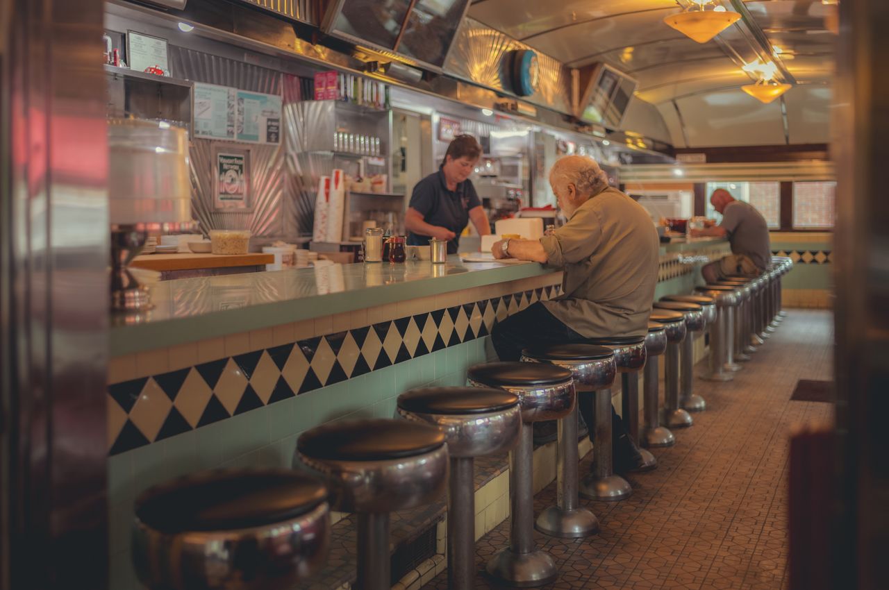 The inside of a classic 50s diner with a lone customer at the counter, surrounded by empty seats.