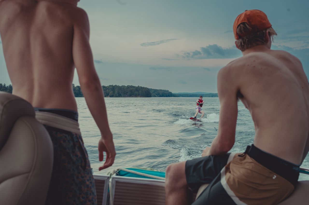 Two men in swim shorts on the back of a boat watching another person wakeboarding behind the boat.