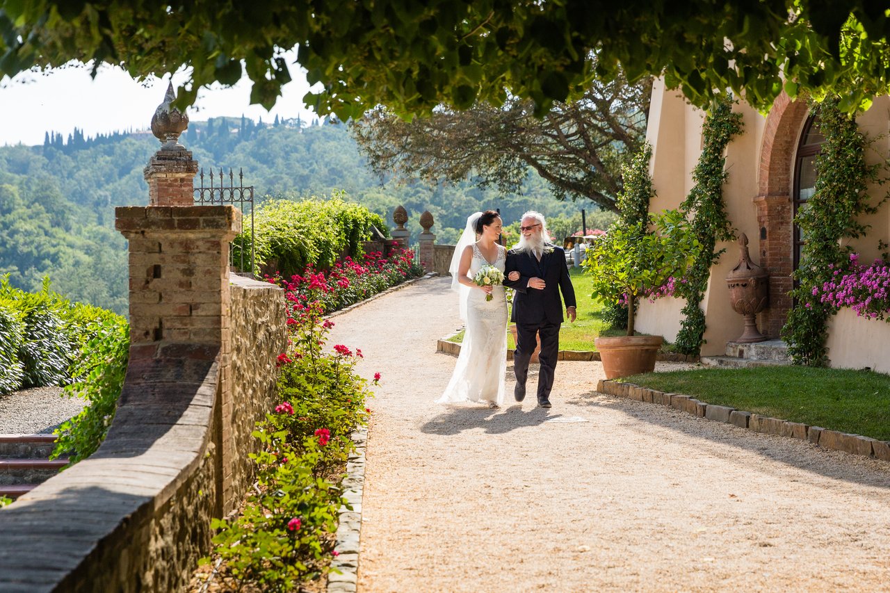 A bride in a white dress walks arm-in-arm with an older man toward the wedding ceremony, holding flowers.