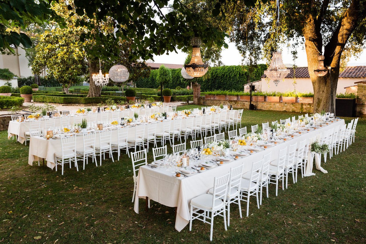 Long outdoor dining tables set for a wedding dinner with white chairs, elegant table settings, and hanging chandeliers.