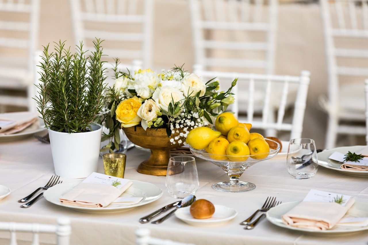 Elegant wedding dinner table with white plates, folded napkins, glassware, and centerpieces of lemons, flowers, and potted herbs.