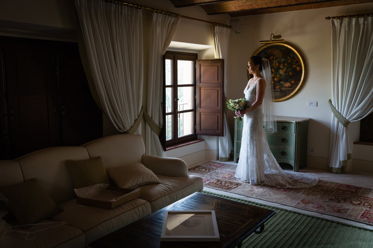 The bride stands by the window in a wedding dress, holding a bouquet and looking outside.