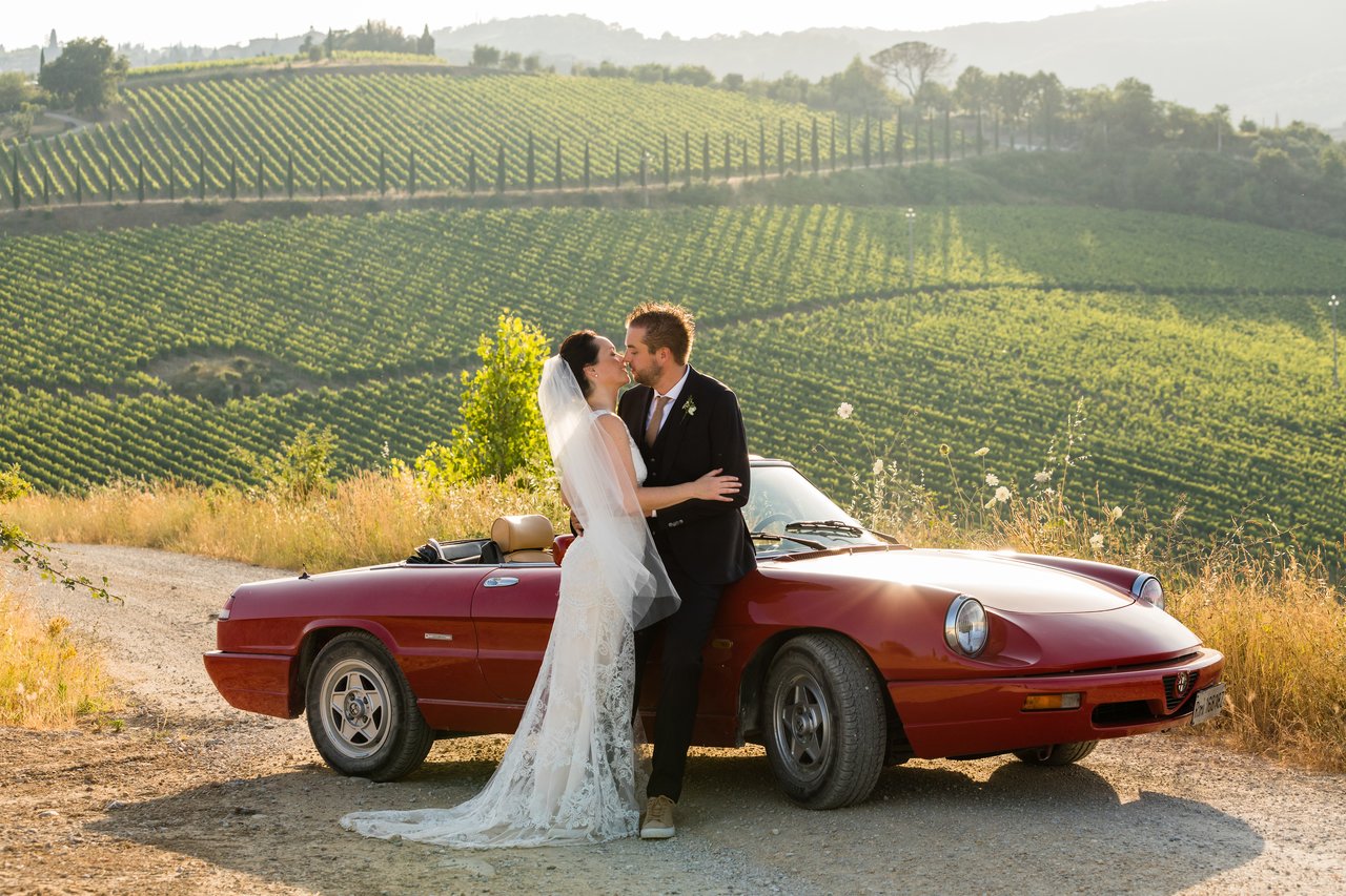 A bride and groom embrace and kiss beside a red convertible, with a vineyard in the background.