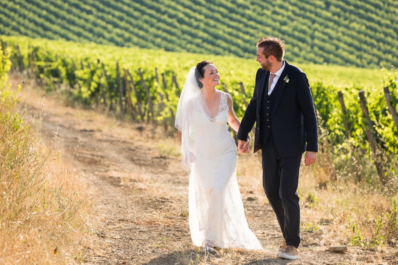 A bride and groom walk hand in hand through a vineyard, smiling at each other.