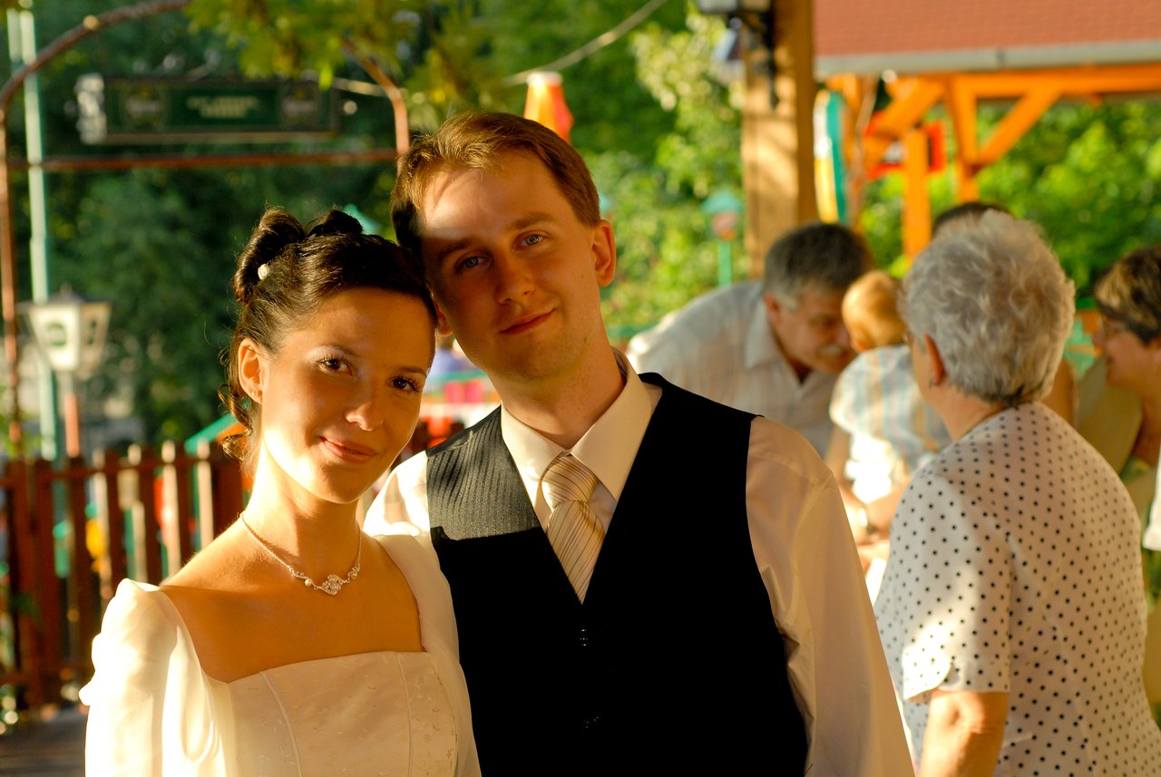 Bride and groom stand close together, smiling at the camera during their wedding celebration with guests in the background.