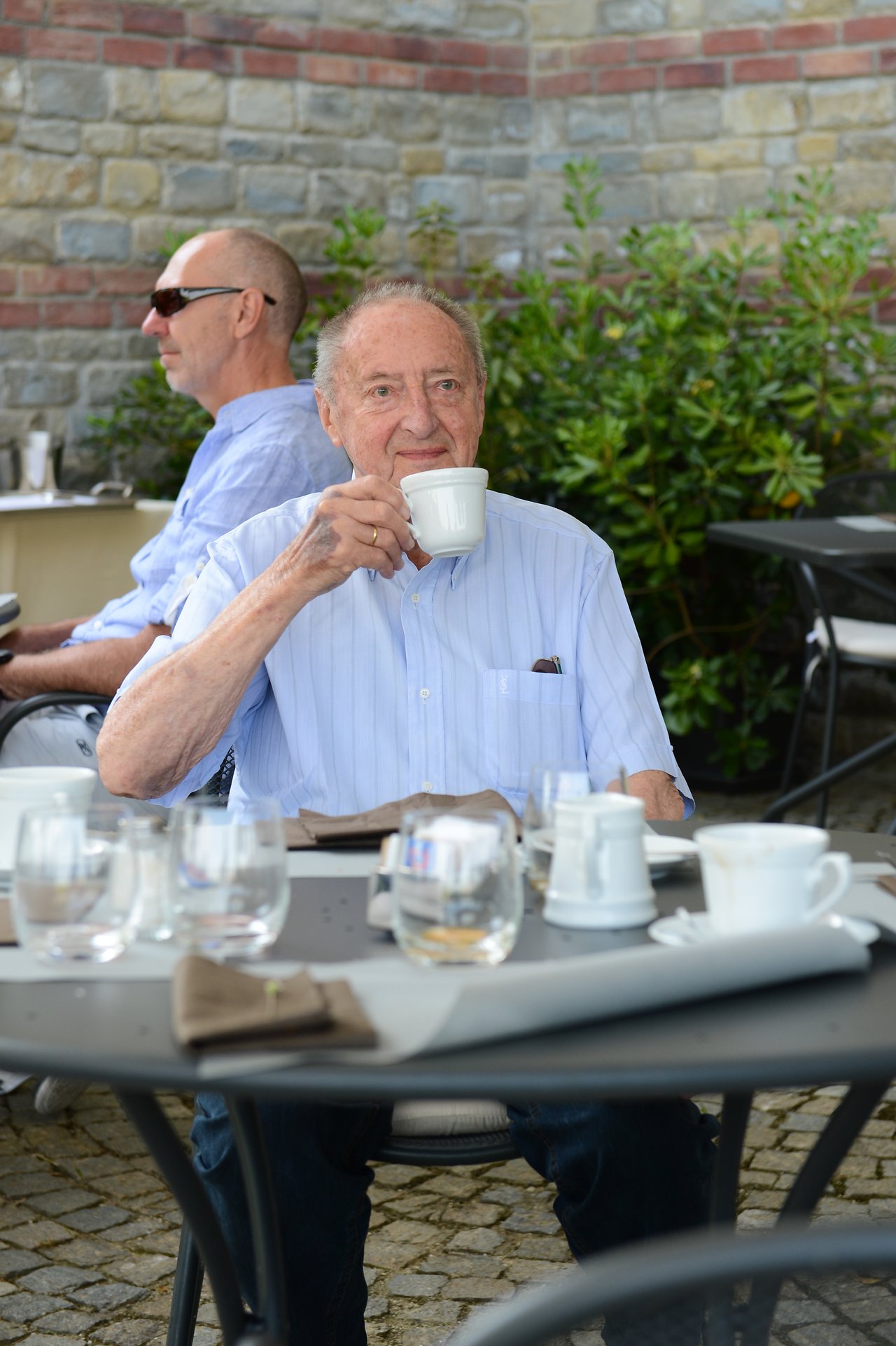 An older man in a light blue shirt drinks coffee at an outdoor table during a wedding gathering.