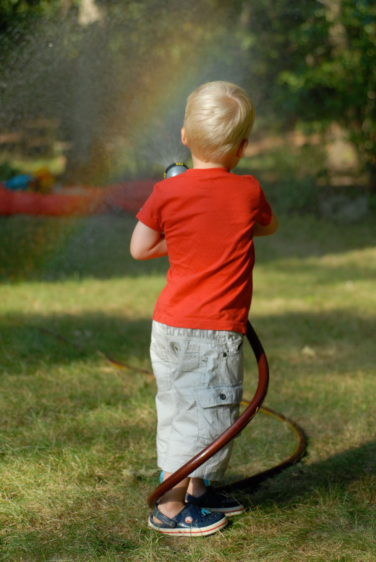A child in a red shirt sprays water from a hose, creating a rainbow in the sunlight.
