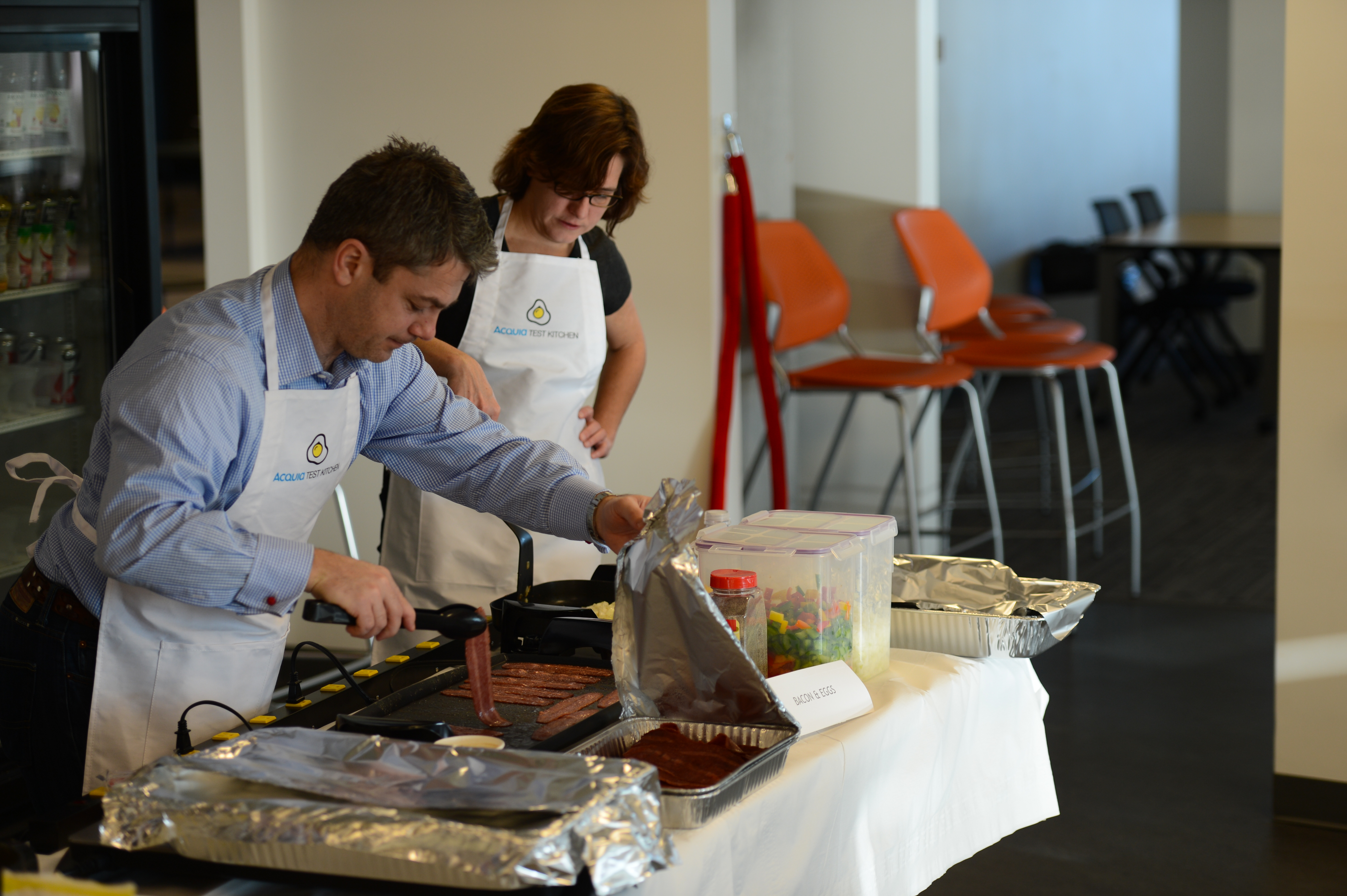 Two people wearing aprons cook breakfast on a griddle at an Acquia event, preparing food on a table.