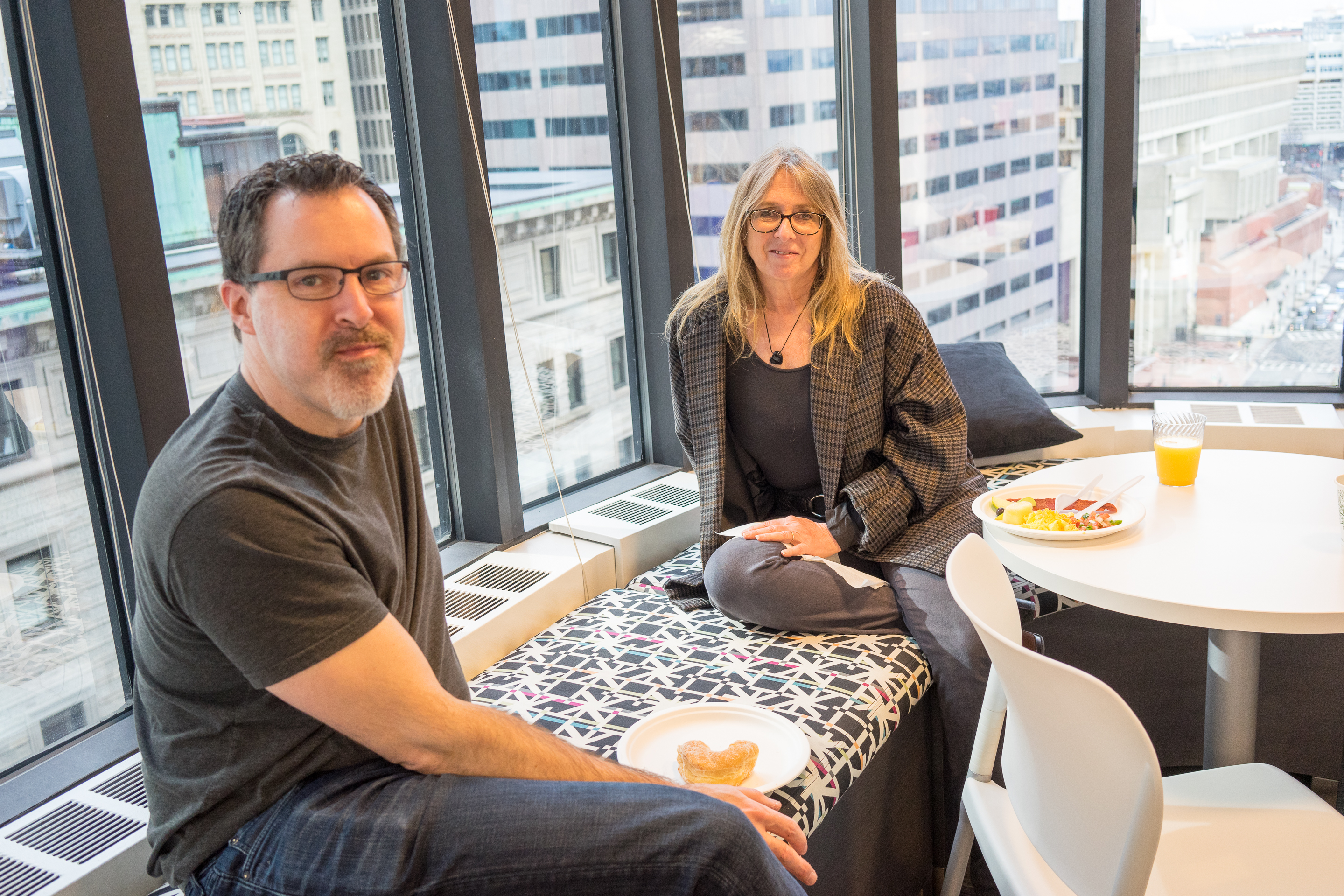 Two people sitting near a window, having breakfast with plates of food and drinks on a table nearby.