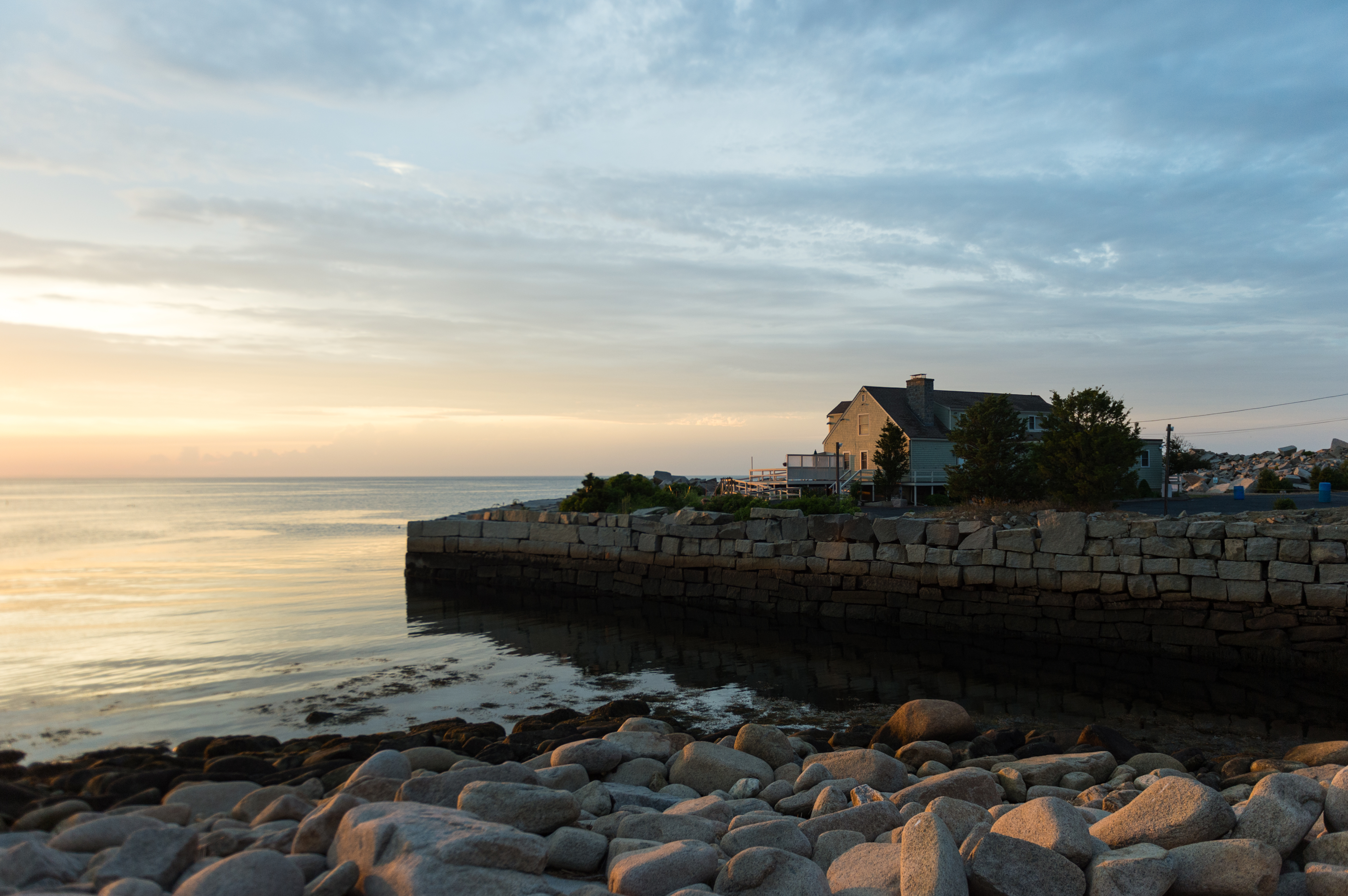 A stone-lined harbor with calm water, a house in the background, and a rocky shoreline at sunset.