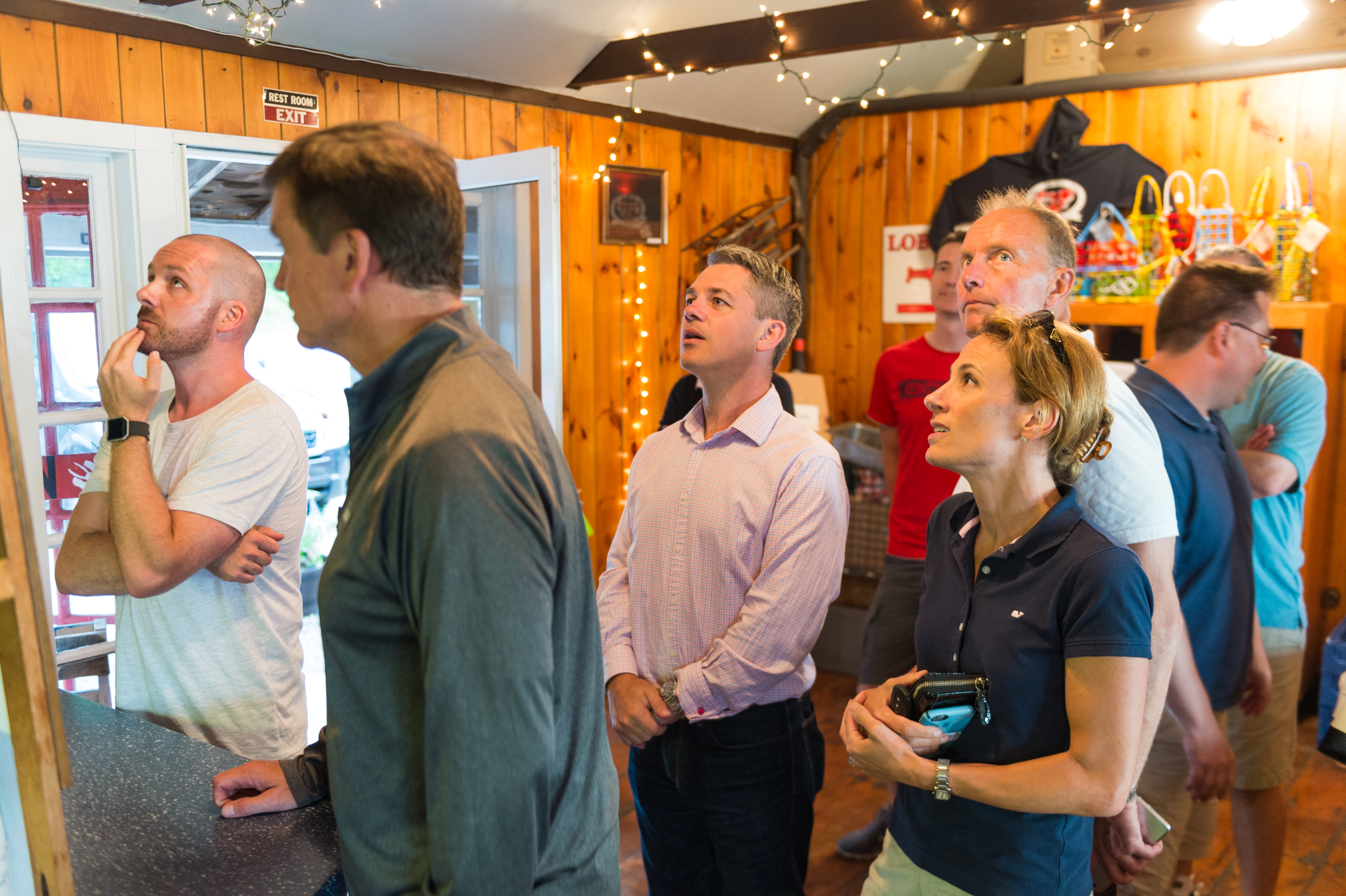 A group of people inside a wooden restaurant look up at a menu, appearing to decide what to order.