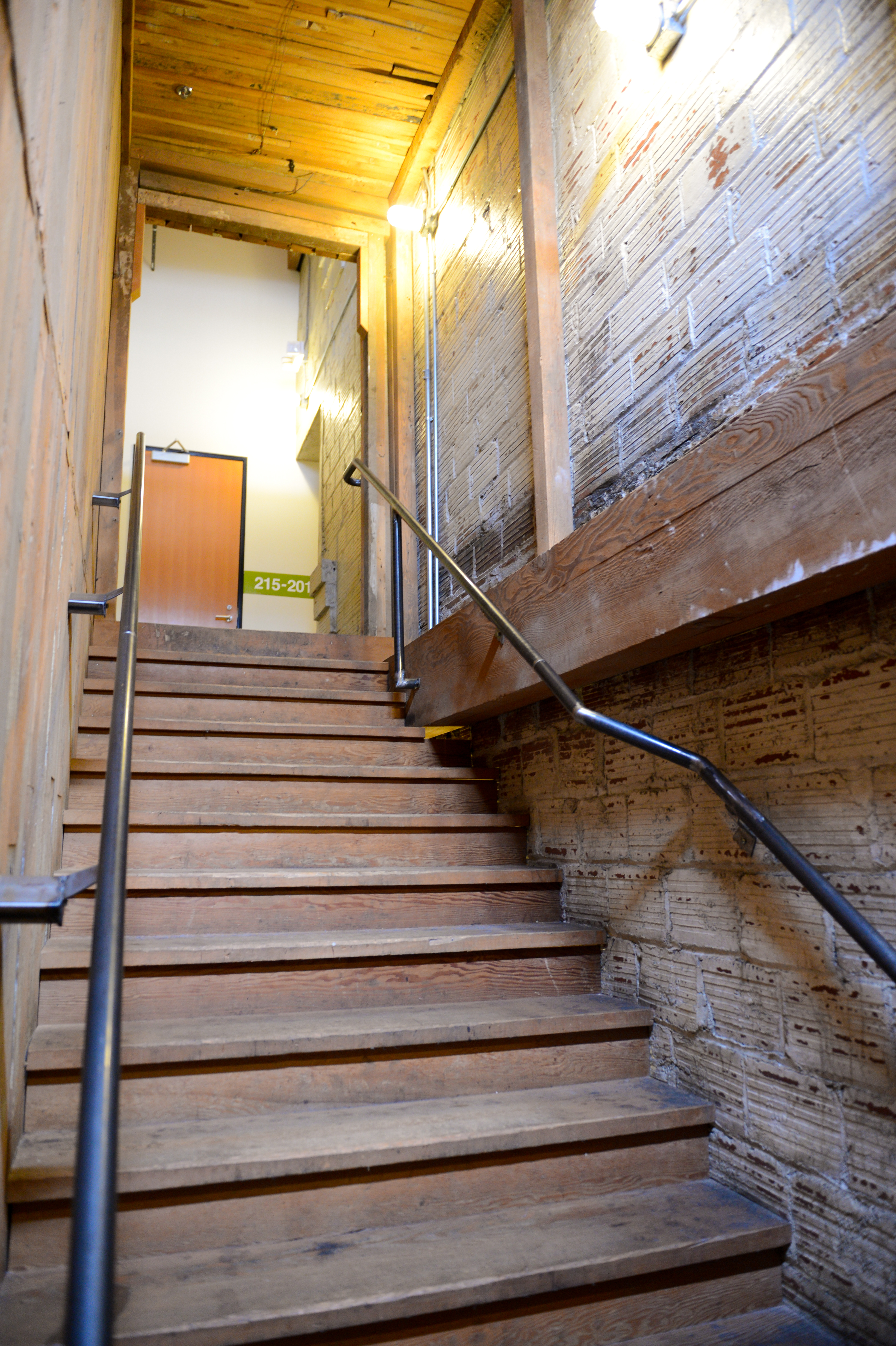 A wooden staircase with metal handrails leads to an office door with a room number sign.