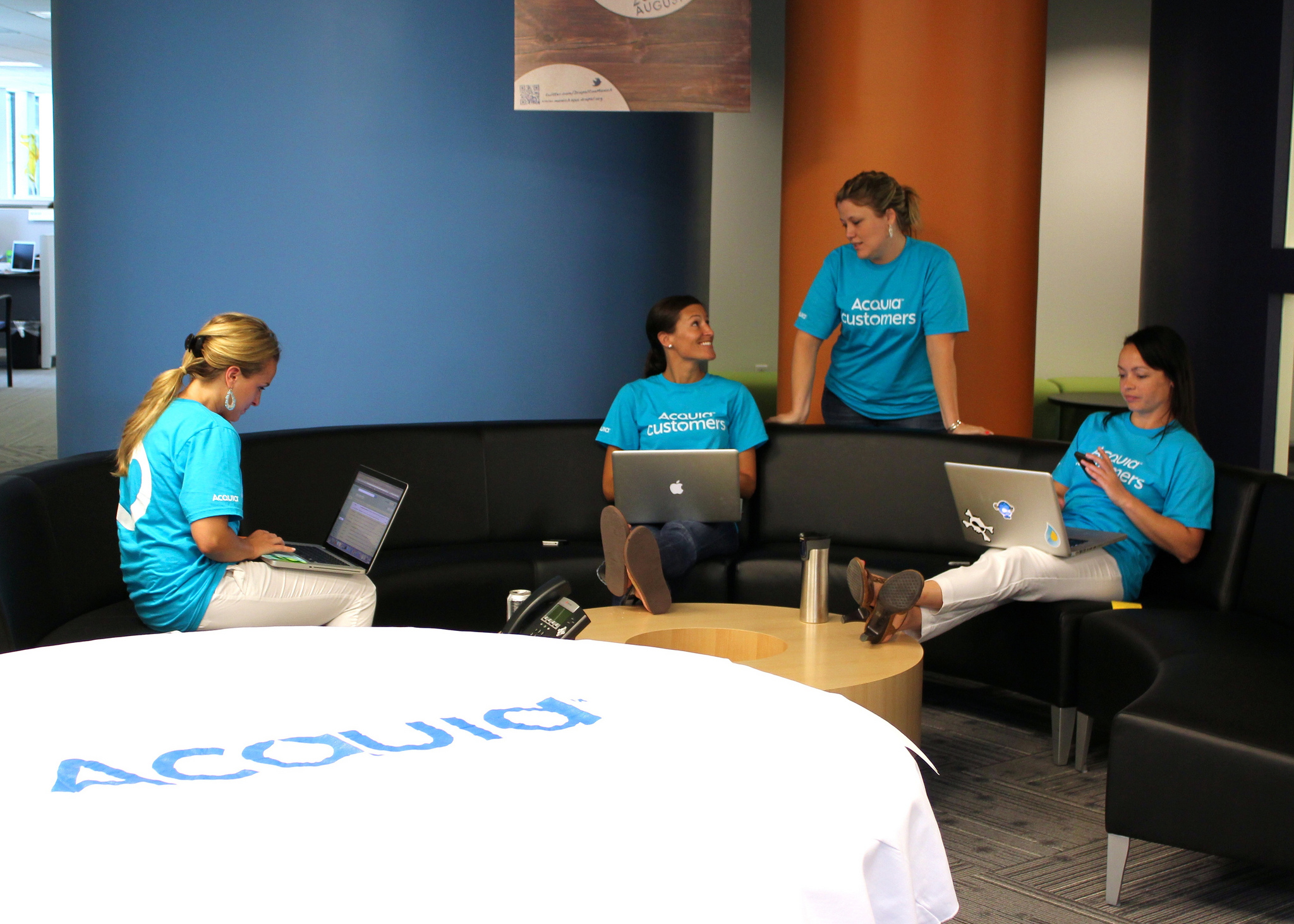 Four people in Acquia shirts sit and stand around a couch, working on laptops and talking during Customer Support Day.